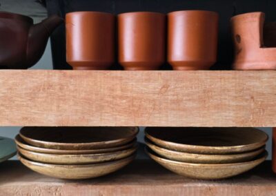 Rustic wooden shelf displaying brown ceramic cups, teapot, and stack of plates.